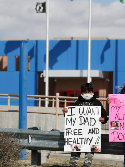 Two children wear masks and hold signs that read "I want my dad free and healthy" and "I want my dad alive not dead!!" as part of a protest calling for the release of detained immigrants in front of the GEO Detention Center in Aurora, Colorado.