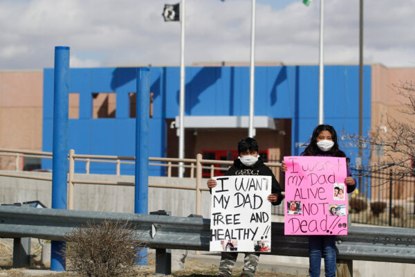 Two children wear masks and hold signs that read "I want my dad free and healthy" and "I want my dad alive not dead!!" as part of a protest calling for the release of detained immigrants in front of the GEO Detention Center in Aurora, Colorado.
