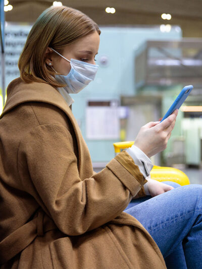 A woman wearing a face mask sits at an airport while scrolling through her phone.
