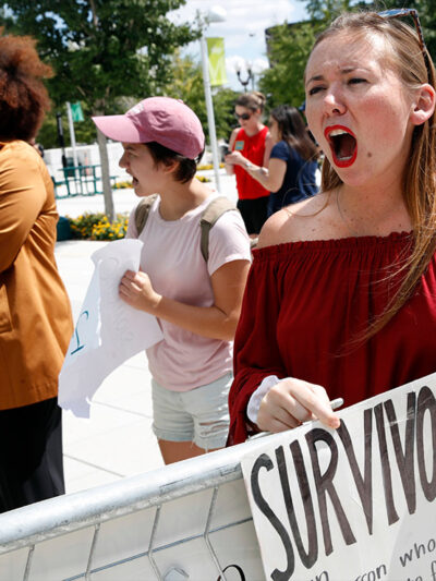 A recent graduate shouts and holds sign that reads "survivor" as Betsey Devos speaks on college campus about changes to Title IX