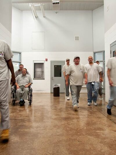 Prisoners are seen walking in a hallway in a prison, one in a wheelchair