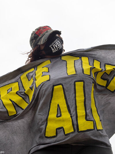 A masked protester is seen wrapped in a sign that says FREE THEM ALL