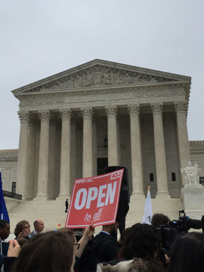 Demonstrators outside the Supreme Court with signs advocating for the rights of LGBT people, including a sign with the text "Open to All" in the center.