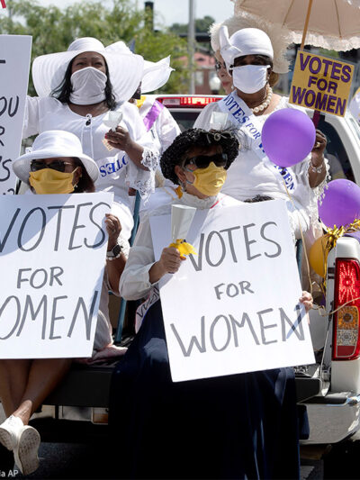Demonstrators in period clothing with signs advocating for women's suffrage.
