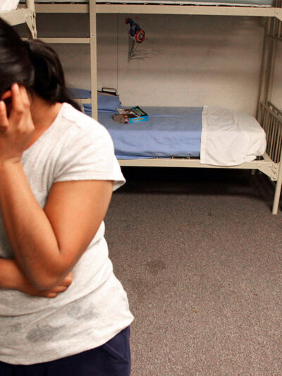A migrant detainee stands facing away from bunks.