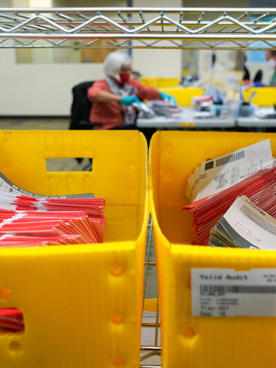 Boxes of vote-by-mail ballot envelopes with workers counting ballots in the background.