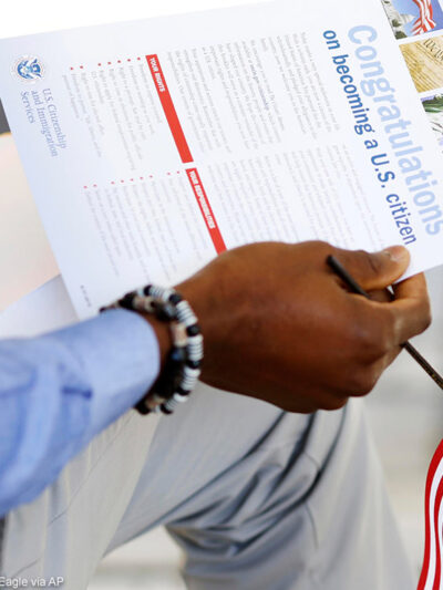 The hand of a seated person holding a miniature U.S. flag and immigration information at a naturalization ceremony.