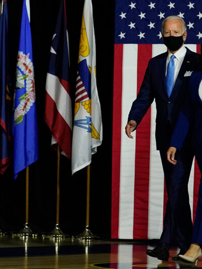 President-Elect Joe Biden and Vice President-Elect Kamala Harris arrive on stage at campaign event with American flag in background.