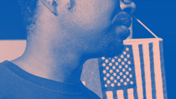 A man wears an "I Voted" stick with the American flag hanging in the background. Image tinted blue and pink.