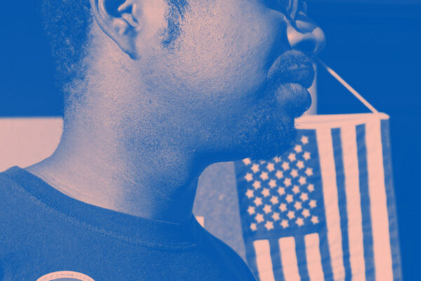A man wears an "I Voted" stick with the American flag hanging in the background. Image tinted blue and pink.