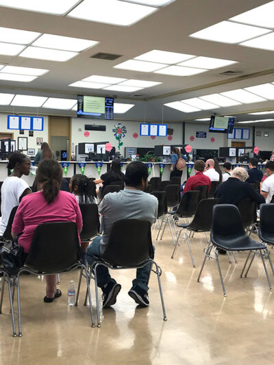 Long rows of chairs with people sitting inside the busy waiting room area of a DMV office.
