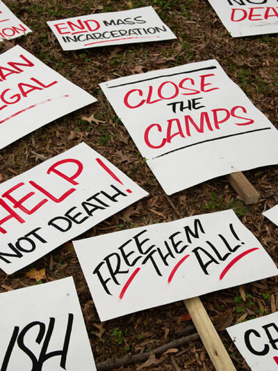 A group of signs on the ground protesting harmful immigration policies, some of which read “No Human Is Illegal,” and “Help Not Death.”