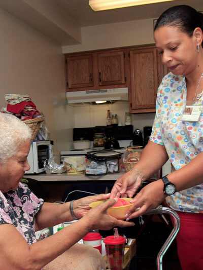 A home health care aide handing her patient a bowl of food.