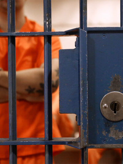 The lock and bars of a prison cell are at the forefront, while a detainee, standing in an orange jumpsuit and with his tattooed arms crossed waits in a cell with other detainees.