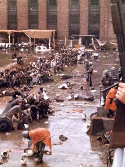 With helmets, gas masks and armed with shotguns, state police overlook the Attica prison yard which is filled with prisoners, face down in mud with their hands behind their heads. Other policemen are in orange ponchos are walking and searching the...