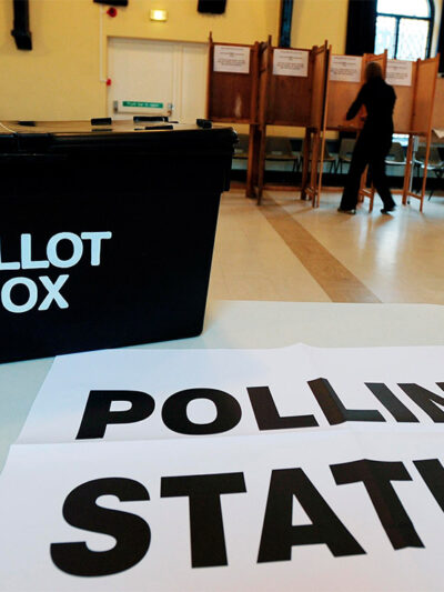In the foreground, a black box with the words "BALLOT BOX" rests on a sign reading "POLLING STATION" at a polling station, while in the background a person in silhouette is at a voting booth.