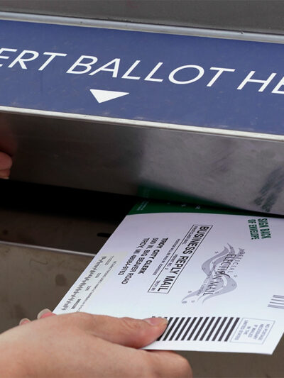 A voter holds the door of a ballot box open with her left hand while inserting her voter ballot with her right hand.