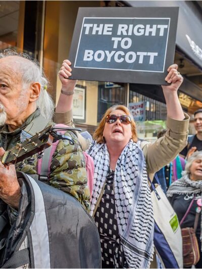 Protestors being led by a man playing a guitar, followed by women holding signs saying" THE RIGHT TO BOYCOTT", and "BOYCOTT UNTIL THE SIEGE ON GAZA ENDS".