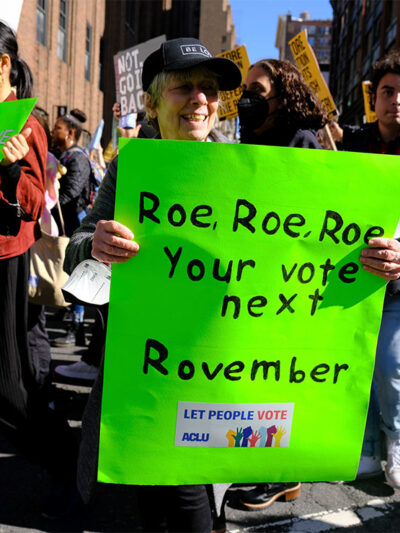 A woman holding a sign reading "Roe, Roe, Roe, Your vote next Rovember / LET PEOPLE VOTE ACLU" poses for the camera as fellow activists walk behind her.