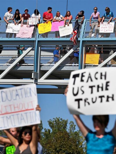In the foreground (at an intersection,) two protesters carry signs with one reading "EDUCATION WITHOUT LIMITATION" and the other "TEACH US THE TRUTH", while in the background, other student demonstrators line an overpass protesting a proposal to...