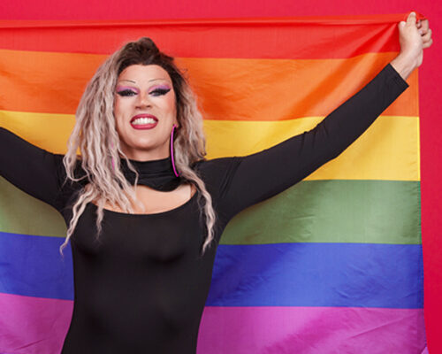 A drag queen holds up a rainbow flag with pride. Show your support by donating to the Drag Defense Fund to protect this freedom of expression.