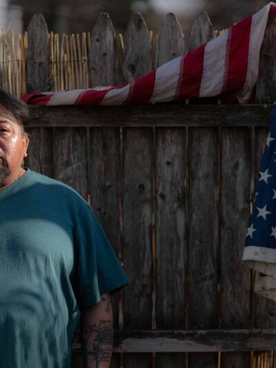Margarito Casta​​ñon Nava looks into the camera as he stands next to an American flag hanging from a wooden fence behind him on his right.