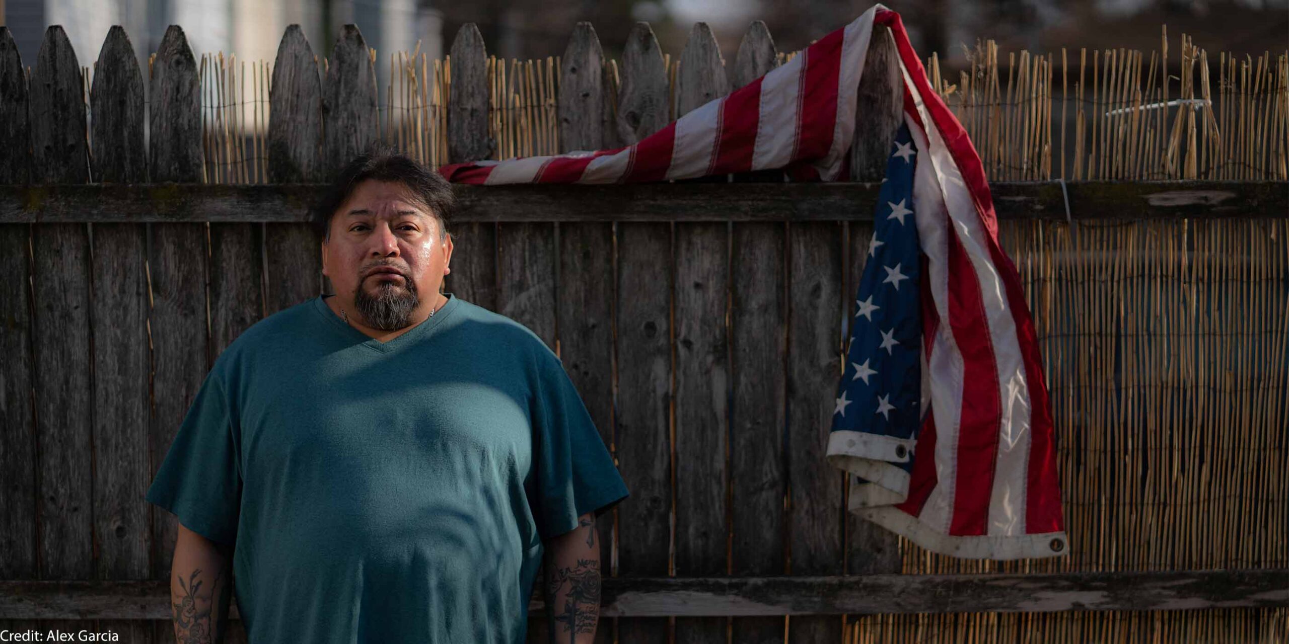 Margarito Castañon Nava looks into the camera as he stands next to an American flag hanging from a wooden fence behind him on his right.