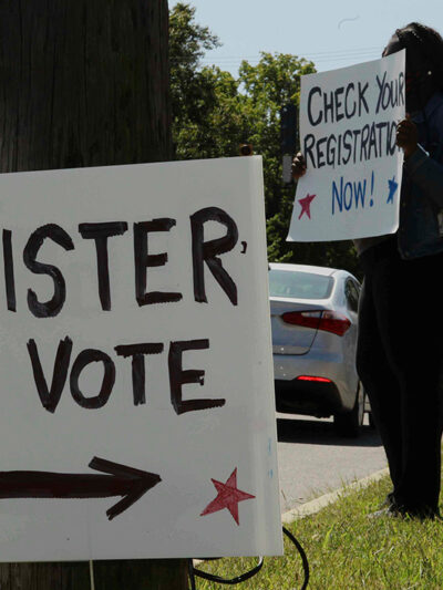 In the foreground, posted on a telephone pole reads a sign "REGISTER TO VOTE", while a woman holds a sign saying "CHECK YOUR REGISTRATION NOW!" is in the background.