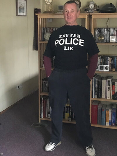 Robert Frese standing in a livingroom wearing a black t-shirt with a message in white lettering reading "EXETER POLICE LIE."