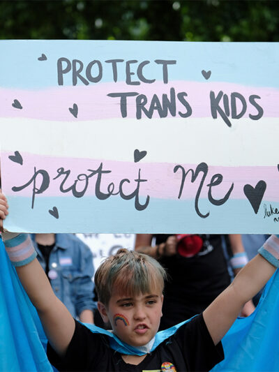A young child holds up a sign reading "PROTECT TRANS KIDS, PROTECT ME - luke, age 9" at a Trans Pride March.