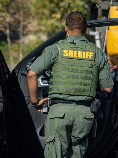 Shot of sheriff (wearing a bulletproof vest) from behind walking towards the open door of police car.