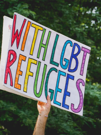 A hand holding up a "With LGBT Refugees" sign.
