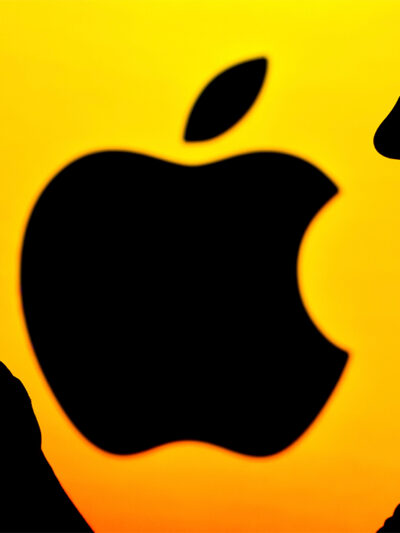 In shadow, a person holds a phone with Apple logo (also in shadow) against a yellow background.
