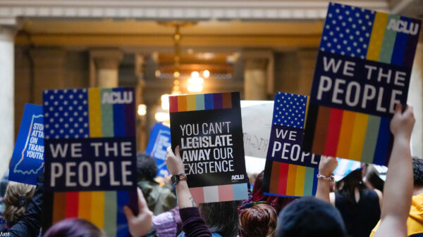 Individuals in a group holding ACLU-branded signs saying "We the People," and "You Can't Legislate Away Our Existence."