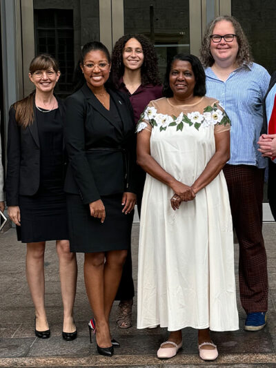 A smiling Leah Watson (a black woman wearing glasses, black business attire and a pin on her left shoulder stands next to a woman wearing a white flower dress wearing glasses and black) gathers for a picture with colleagues.
