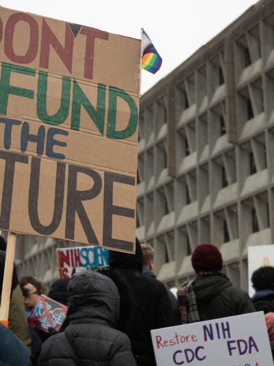 People attend a rally against the Trump administration's 90-day funding freeze and job cuts at health agencies in Washington, DC, hold up a large sign written in red, green and black on cardboard that reads " DON'T DEFUND THE FUTURE."
