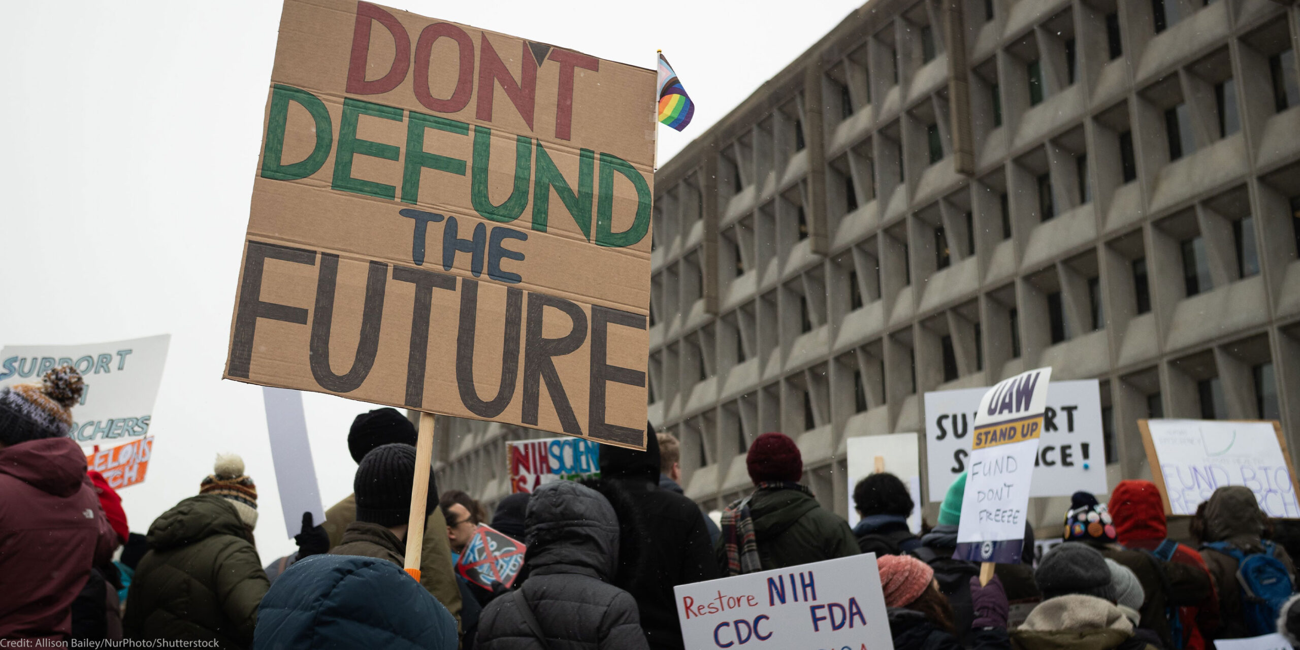 People attend a rally against the Trump administration's 90-day funding freeze and job cuts at health agencies in Washington, DC, hold up a large sign written in red, green and black on cardboard that reads " DON'T DEFUND THE FUTURE."