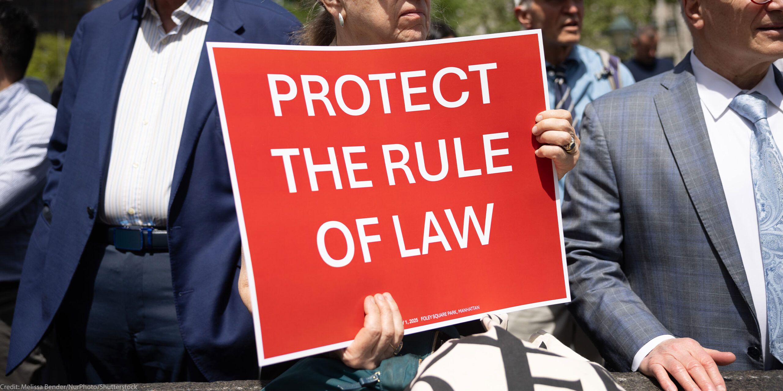 At the Lawyers Rally In Lower Manhattan To Demand Respect For The Rule Of Law, a demonstrator holds up a sign with white lettering on a red background that reds, "PROTECT THE RULE OF LAW".