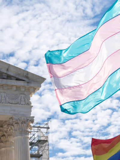 The Trans and LGBTQ+ flags waving in the wind in front of the Supreme Court Building in DC.