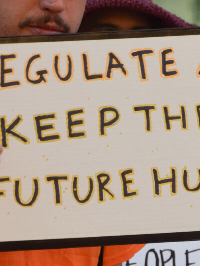 The camera focuses on a sign held by a demonstrator that reads, "REGULATE AI KEEP THE FUTURE HUMAN."