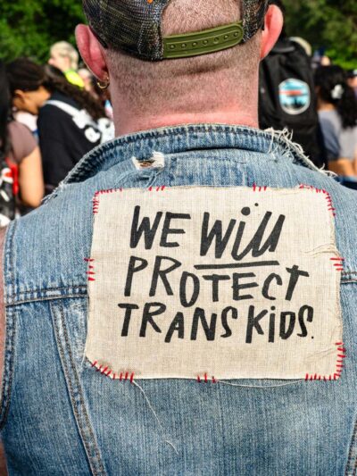At a trans rally, person (wearing a denim vest and with their back to the camera) displays the sign stitched to the vest that reads "WE WILL PROTECT TRANS KIDS."