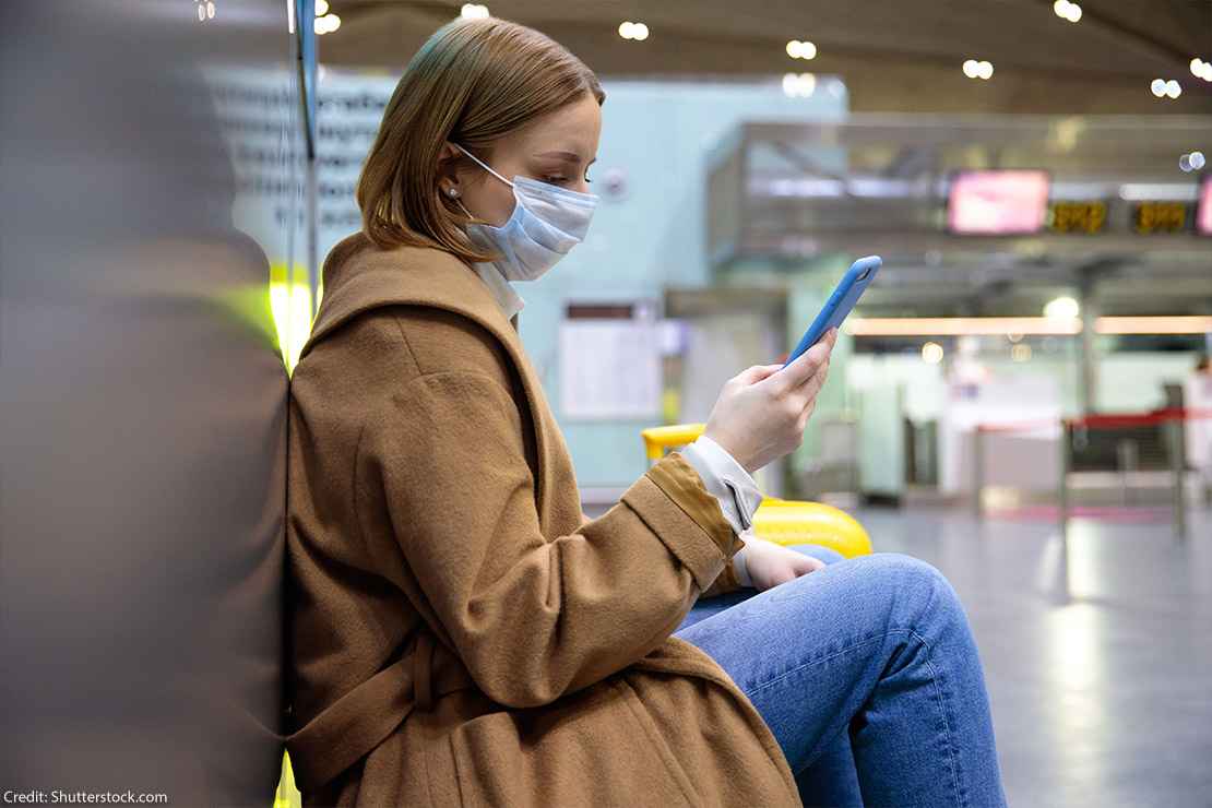 A woman wearing a face mask sits at an airport while scrolling through her phone.