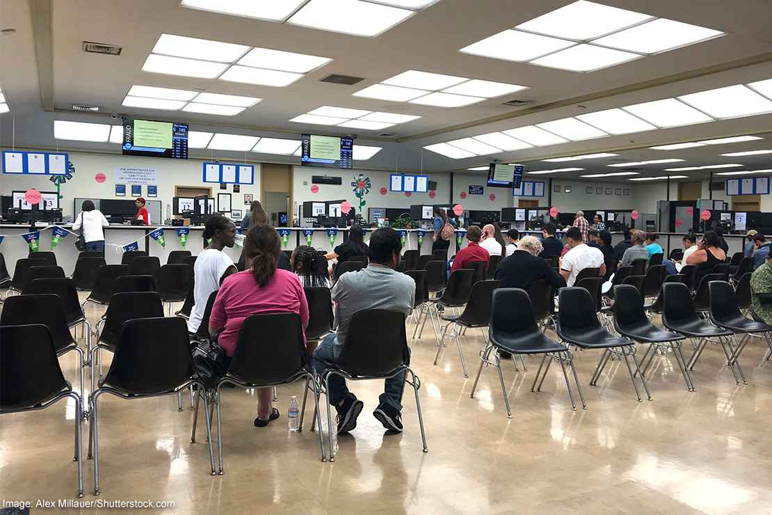 Long rows of chairs with people sitting inside the busy waiting room area of a DMV office.