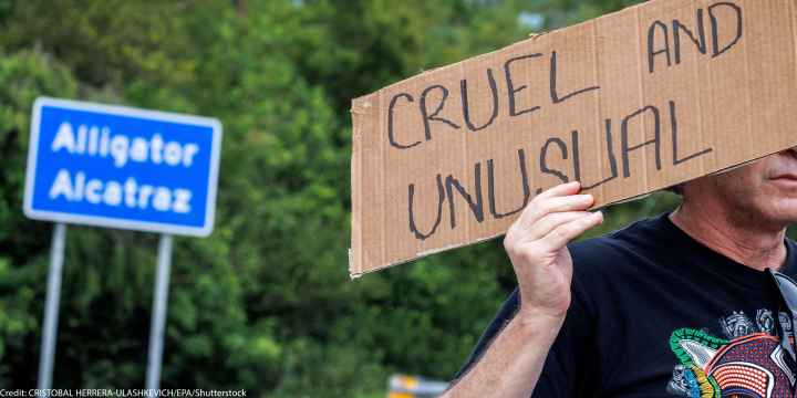 A road sign that says Alligator Alcatraz and an individual in front of it holding a sign that says "Cruel and Unusual."