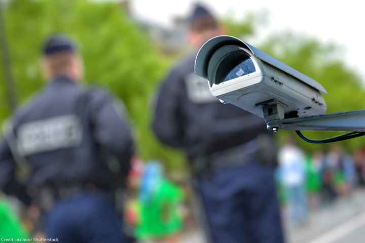 A security CCTV camera or surveillance system with police officers in the blurry background.