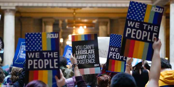 Individuals in a group holding ACLU-branded signs saying "We the People," and "You Can't Legislate Away Our Existence."