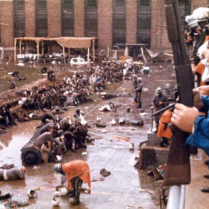 With helmets, gas masks and armed with shotguns, state police overlook the Attica prison yard which is filled with prisoners, face down in mud with their hands behind their heads. Other policemen are in orange ponchos are walking and searching the...