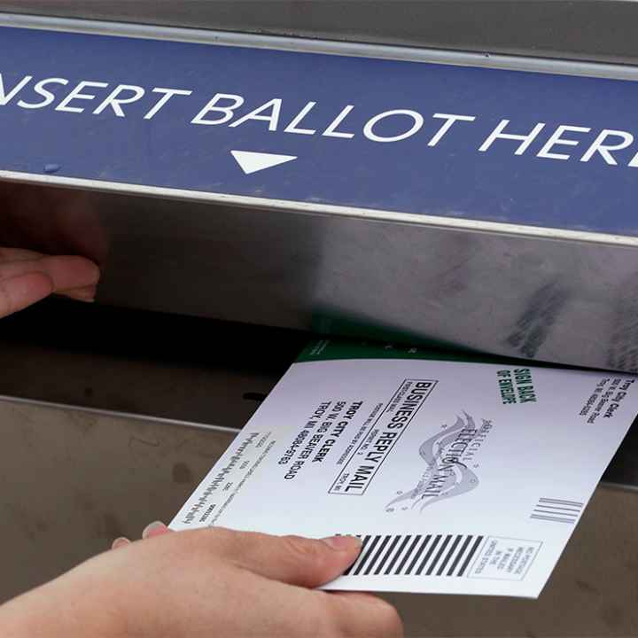 A voter holds the door of a ballot box open with her left hand while inserting her voter ballot with her right hand.