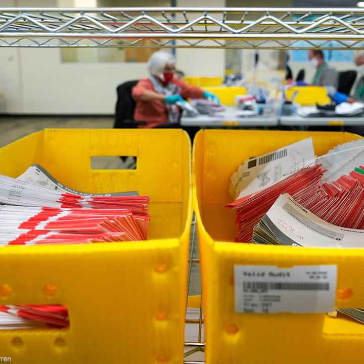 Boxes of vote-by-mail ballot envelopes with workers counting ballots in the background.
