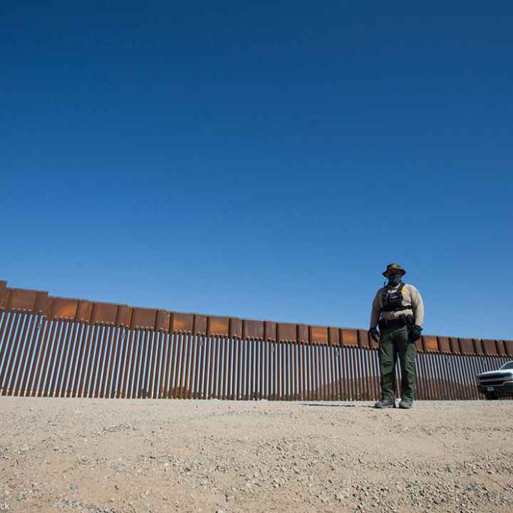 A uniformed individual in front of a portion of U.S., Mexico border wall.